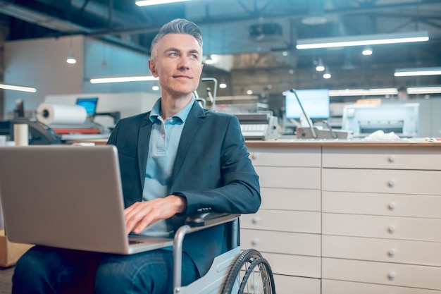 Positive Einstellung. Lächelnder junger erwachsener Mann mit Laptop sitzt im Rollstuhl und schaut nachdenklich zur Seite am Arbeitsplatz im Büro