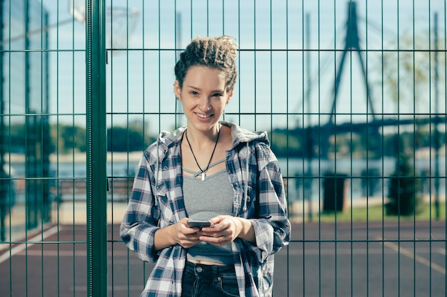 Positive entspannte junge Frau, die modernes Gerät hält und lächelt, während sie neben dem Maschendrahtzaun steht