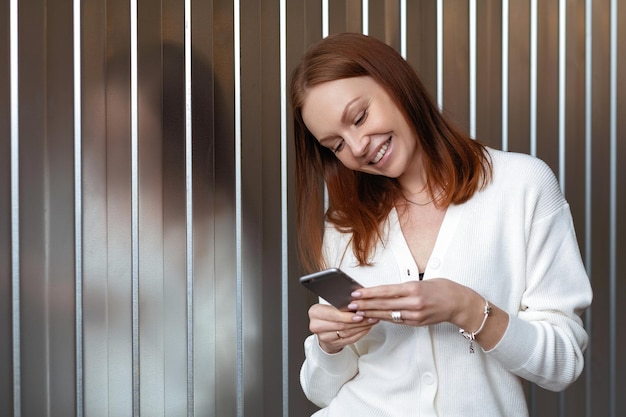 Positive, erfolgreiche europäische Wirtschaftswissenschaftlerin macht Geldzahlung auf Website-Posen mit Handy in weißem Outfit, lächelt glücklich, sendet Textnachricht, genießt Online-Kommunikation