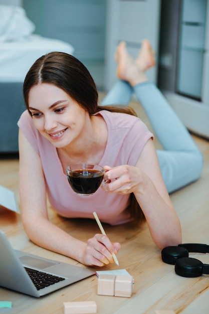 Positive Frau, die auf den Laptop-Bildschirm schaut, während sie mit einer Tasse in der Hand auf dem Boden im Schlafzimmer liegt