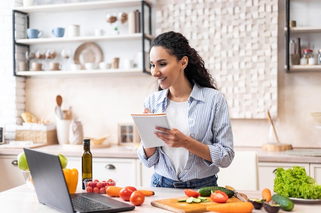 Positive Frau, die ein neues Rezept schreibt, während sie sich ein Video auf dem Laptop ansieht, das in einem modernen Kücheninterieur kocht