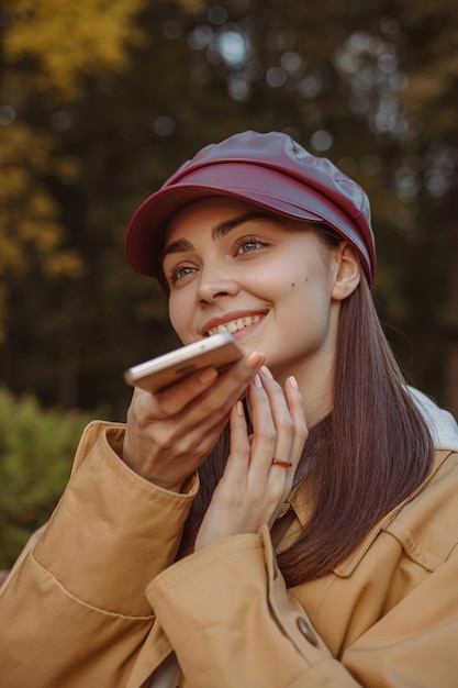 Positive Frau, die Sprachnachrichten auf dem Smartphone aufzeichnet, während sie über soziale Medien im Park kommuniziert