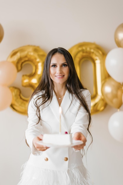 Positive Frau feiert Jubiläum in stilvollem Federkleid, das zu Hause mit Geburtstagskerzenkuchen um aufgeblasene Luftballons posiert
