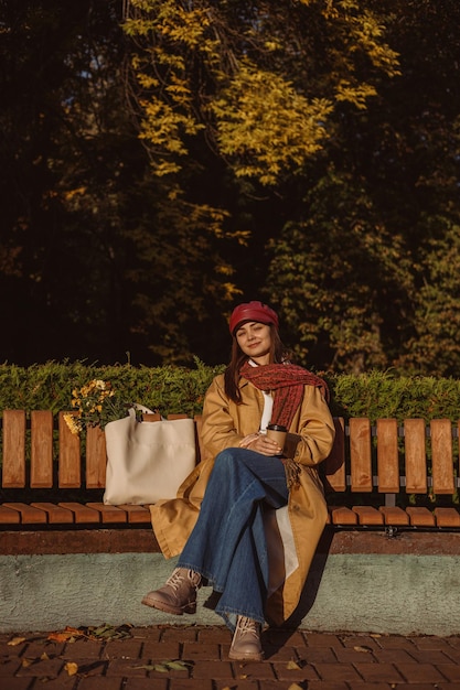 Positive Frau in Graben und Mütze mit Kaffeetasse zum Mitnehmen, die sich auf der Bank im Herbstpark entspannt