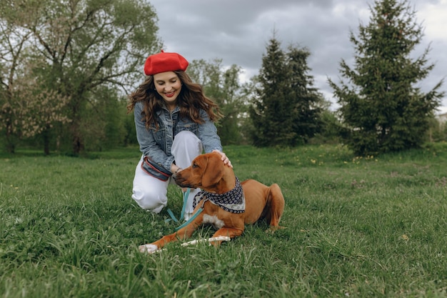 Positive Frau in roter Baskenmütze, die braunen Hund streichelt, während sie zusammen auf einer Wiese in der Natur sitzt