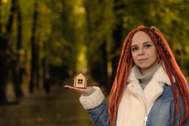 Positive Frau mit Dreadlocks, die ein kleines Holzhaus im Waldpark hält. Frau mit Spielzeughaus, die lächelnd in die Kamera blickt. Konzept des Kaufs eines neuen Wohnungsdachs an einem ökologischen Ort