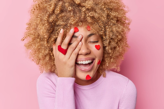 Positive frau mit lockigem haar macht gesicht palme mit kleinen roten herzen geschmückt hält die augen geschlossen lächelt zahnhaft isoliert über rosa hintergrund fröhliches weibliches modell, das verliebt ist, hat romantische stimmung