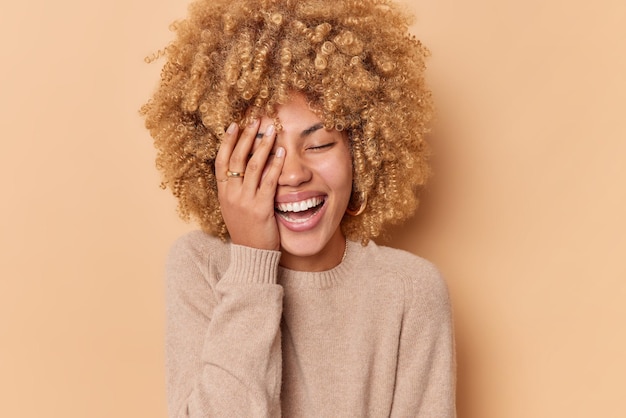 Positive Frau mit lockigen blonden Haaren lässt das Gesicht mit der Handfläche lächeln, schließt positiv die Augen und drückt aufrichtige Emotionen aus