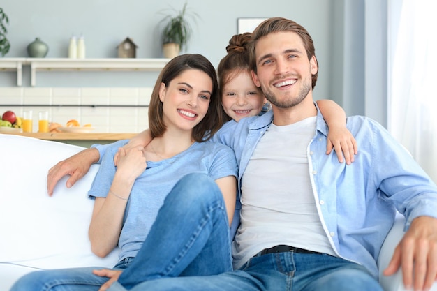 Positive, freundliche junge Eltern mit lächelnder kleiner Tochter, die zusammen auf dem Sofa sitzen, während sie sich am Wochenende zu Hause entspannen.