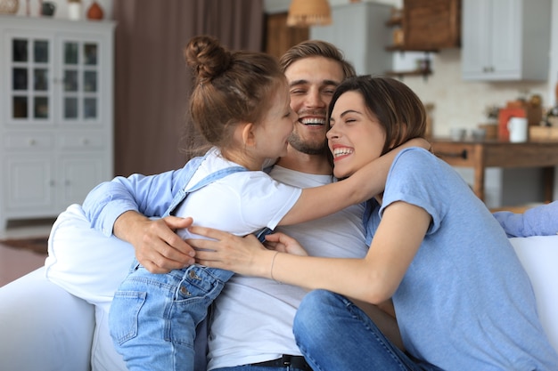 Positive freundliche junge Eltern mit lächelnder kleiner Tochter, die zusammen auf dem Sofa sitzt und sich am Wochenende zu Hause entspannt.