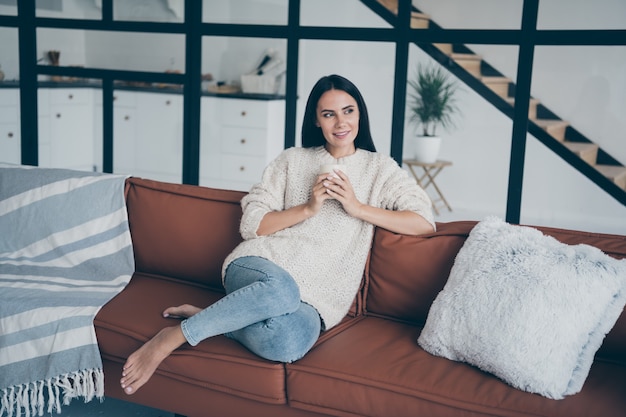 Positive fröhliche Frau sitzen auf braunem Leder Diwan halten Tasse mit Latte Cappuccino starren Fenster fühlen sich inspiriert Fantasie verträumt im Haus drinnen