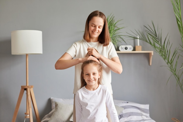 Positive, fröhliche Mutter, die ihrer Tochter eine Frisur macht, während sie im Schlafzimmer auf dem Bett sitzt und dem Kind einen Pferdeschwanz macht, bevor sie morgens zur Schule geht