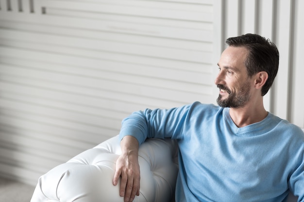 Positive Gefühle. Netter glücklicher bärtiger Mann, der auf dem Sofa sitzt und lächelt, während er eine wunderbare Stimmung hat