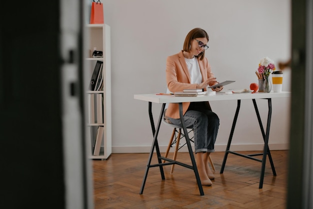 Positive Geschäftsfrau in stylischem Outfit mit Lächeln, die im Tablet arbeitet und am hellen Desktop sitzt