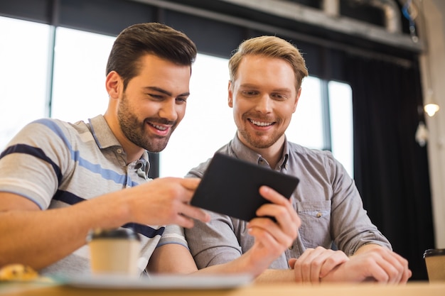 Positive glückliche freudige Männer, die zusammen sitzen und den Tablettbildschirm betrachten, während sie im Team arbeiten