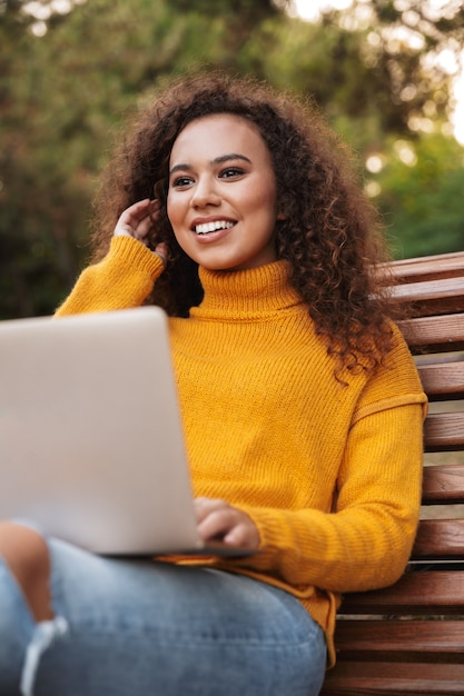 positive glückliche schöne junge lockige Frau sitzen im Park im Freien mit Laptop-Computer.