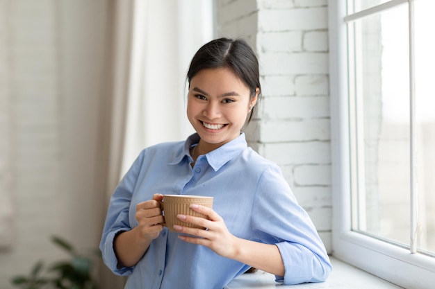 Positive hübsche asiatische Frau, die am Fenster steht und Kaffee trinkt