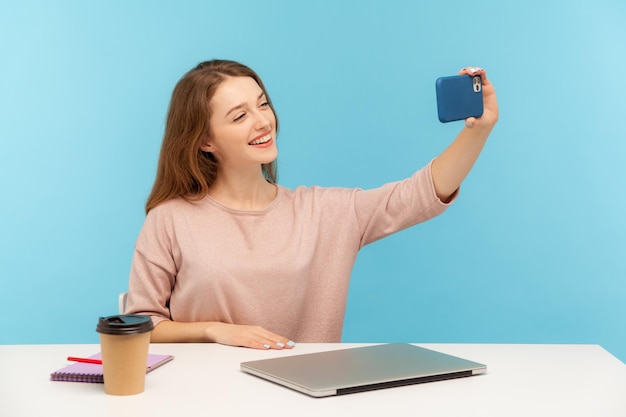 Positive hübsche Frau, Büroangestellte oder Bloggerin, die am Arbeitsplatz sitzt und ein Selfie mit dem Handy macht, bei Videoanrufen freundlich lächelt, mit dem Handy streamt. Studioaufnahme im Innenbereich, isoliert