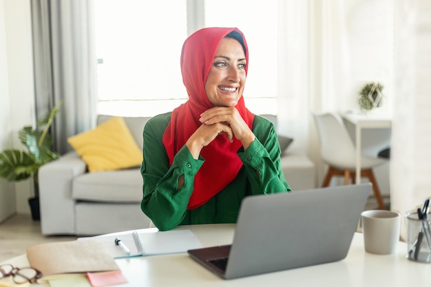 Positive islamische frau im hijab, die zu hause am schreibtisch mit laptop sitzt und über etwas nachdenkt und lächelt