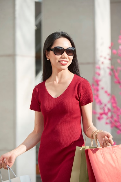 Positive junge Asiatin in Sonnenbrille und rotem Kleid mit Einkaufstüten beim Gehen im Einkaufszentrum