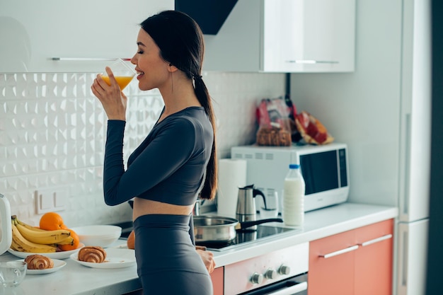 Positive junge Dame, die in der Küche steht und lächelt, während sie Orangensaft trinkt