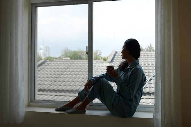 Positive junge Frau, die auf der Fensterbank sitzt, Morgenkaffee trinkt und nach draußen schaut