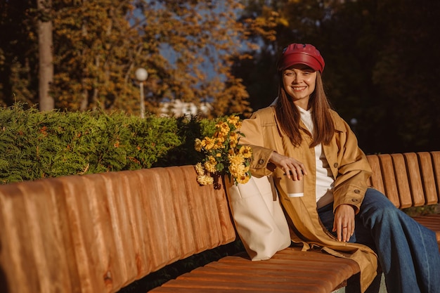 Positive junge Frau im Trenchcoat mit Pappbecher Kaffee zum Sitzen auf der Bank mit Blumen in der Tasche