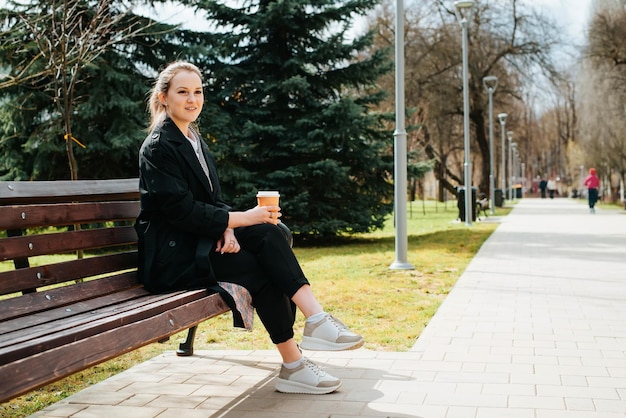 Positive junge Frau in stylischem Mantel, die auf einer Parkbank ruht. Seitenansicht charmante lächelnde Frau mit einer Tasse Kaffee, die weg vom Kopierraum schaut