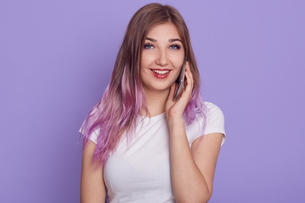 Positive junge Frau mit angenehmem Aussehen und violettem Haar, trägt weißes T-Shirt, spricht über Handy, schaut lächelnd direkt in die Kamera, einzeln auf lila Hintergrund.