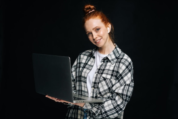 Positive junge Frau mit drahtlosen Kopfhörern, Laptop-Computer in der Hand und Blick in die Kamera