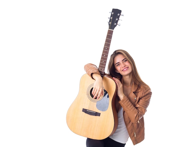 Positive junge Frau mit langen Haaren in brauner Jacke, die eine Gitarre auf weißem Hintergrund im Studio hält