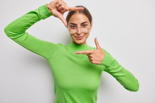 Positive junge Frau sucht perfekte Winkelperspektive oder Inspiration macht Rahmengeste trägt große optische Brillen und grüner Rollkragenpullover macht gute Aufnahmen isoliert über grauer Wand