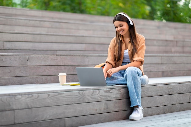 Positive junge Hipsterdame mit Laptop und Kopfhörern im Freien