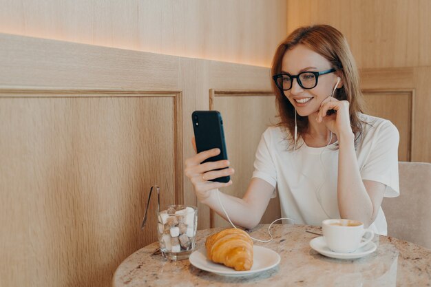 Positive junge rothaarige Bloggerin hat Video-Chat mit Followern und hält modernes Handy