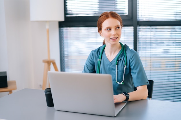 Positive junge Ärztin in blauer medizinischer Uniform, die auf der Laptop-Tastatur am Schreibtisch sitzt