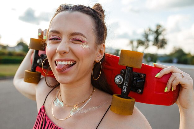 Positive junge Teenager-Skaterin mit Skateboard, die Zunge zeigt und lustiges Gesicht macht