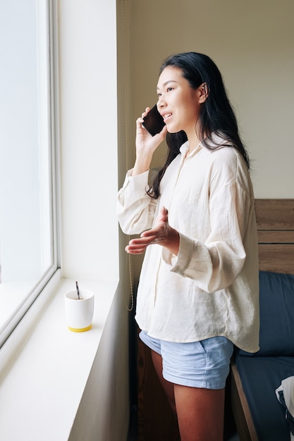 Positive junge Vietnamesin, die in ihrem Schlafzimmer am Fenster steht und mit einem Freund telefoniert?