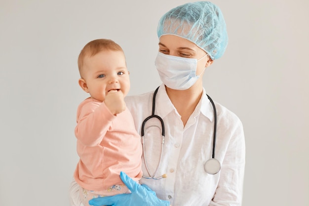 Positive lächelnde junge erwachsene weibliche Kinderärztin, die medizinische Uniform trägt, Säuglingsbaby hält, die Gesundheit des Kindes in der Klinik untersucht, monatliche Untersuchung von Neugeborenen.