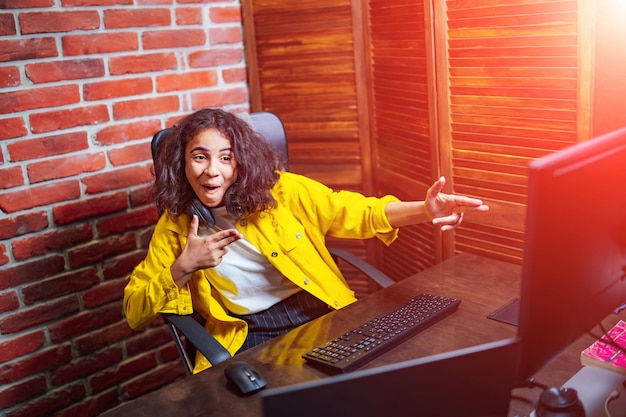 Positive menschliche Emotionen Porträt eines glücklichen, emotionalen Teenager-Mädchens mit lockigem Haar, das mit den Fingern auf dem halb umgedrehten Backsteinmauerhintergrund des Computers angezeigt wird