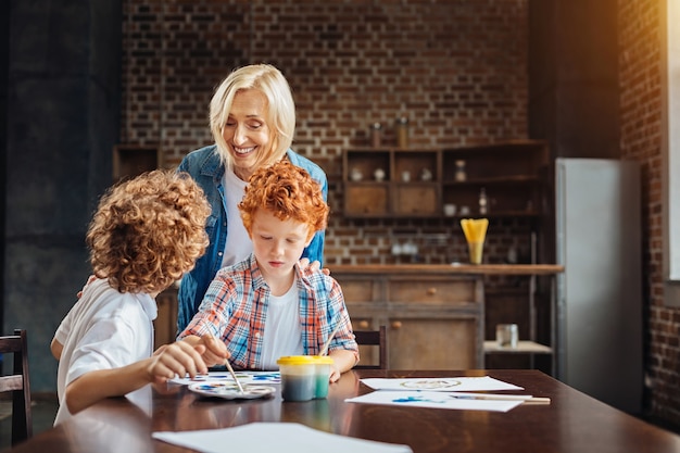 positive reife Frau, die sich ihren Enkelkindern anschließt, die mit Aquarellen malen und breit grinsen, während einer von ihnen hört, der ihr eine Geschichte erzählt.