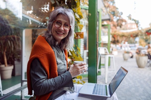 Positive reife Frau mit Brille und Laptop hält eine Tasse in der Nähe des Cafés