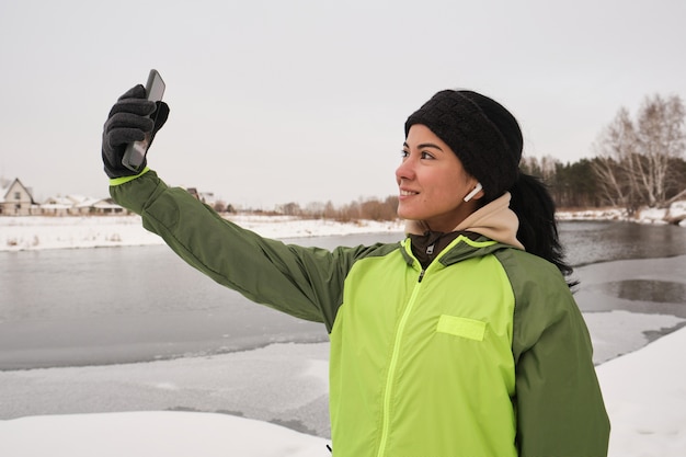 Positive schöne Frau in den Ohrstöpseln, die auf Winterufer stehen und sich auf Smartphone-Kamera fotografieren