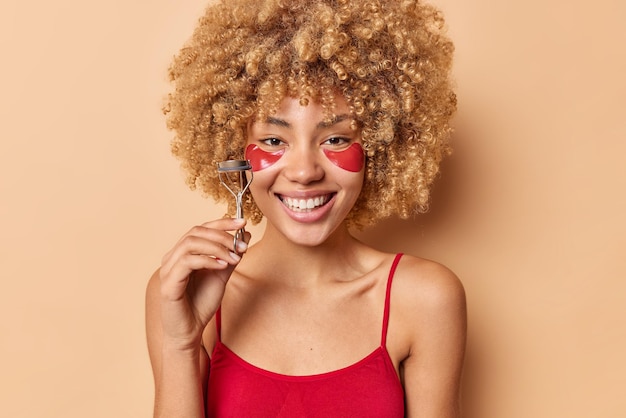 Positive schöne frau mit lockigem haar in lässigem t-shirt hält wimpernwickler, die make-up auftragen wird, trägt hydrogelflecken unter den augen und ist gut gelaunt isoliert über beigem hintergrund