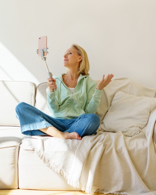 Positive Seniorin, die Selfie auf dem Smartphone macht, während sie sich zu Hause auf dem Sofa entspannt