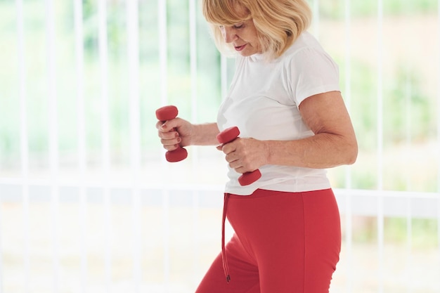 Positive Seniorin in sportlicher Kleidung drinnen tagsüber, die Übungen mit Hanteln macht