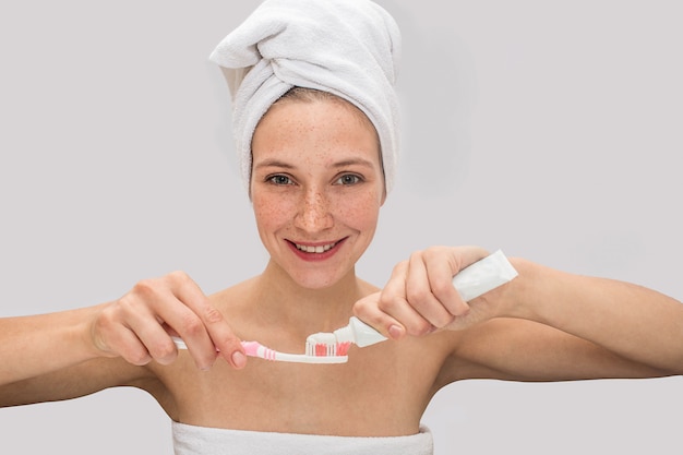 Positive sommersprossige junge Frau schaut und lächelt. Sie gibt Zahnpasta auf die Zahnbürste. Modell hat weiße Handtücher um ihren Körper und Haare.