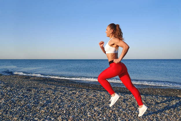Positive sportliche Frau an einem Sommermorgen, der am Strand in den roten Leggings auf dem Hintergrund der Seeküste joggt