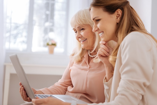 Positive Stimmung. Schöne entzückende nette Frau, die ihr Kinn hält und auf den Laptopbildschirm schaut, während sie zusammen mit ihrer Mutter sitzt