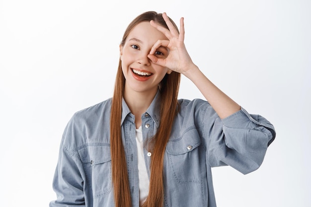 Positive Teenager-Mädchen Studentin sagen okay Show OK Geste auf dem Auge und lächelnd sagen ja genehmigen oder empfehlen etwas Cooles Stehen erfreut vor weißem Hintergrund