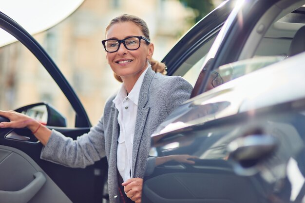 Positive und erfolgreiche Geschäftsfrau mittleren Alters in klassischer Kleidung, die aus ihrem modernen Auto steigt